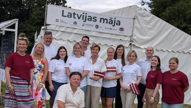 Cilvēku grupa pozē kopbildei pie “Latvijas mājas” telts āra festivālā