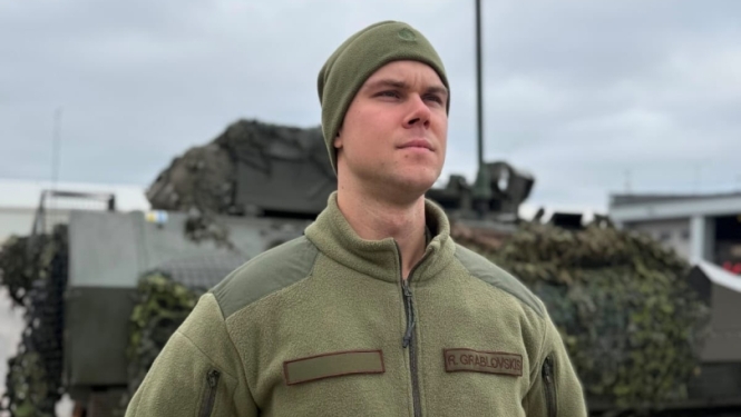 Vīrietis militārajā apģērbā un cepurē stāv pie maskēta bruņutransportiera