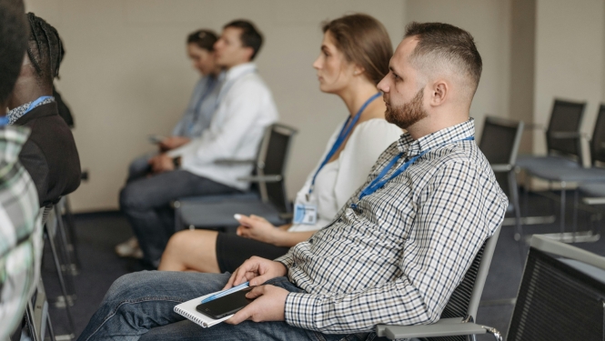 Cilvēki sēž seminārā vai konferencē, klausoties prezentāciju telpā