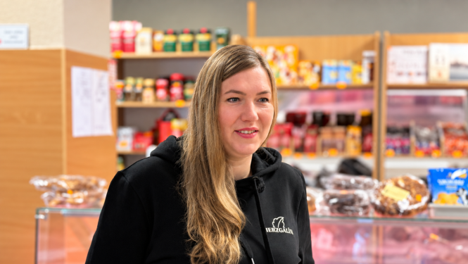 Sieviete pārtikas veikala telpā stāv pie vitrīnas ar produkciju un plauktiem fonā