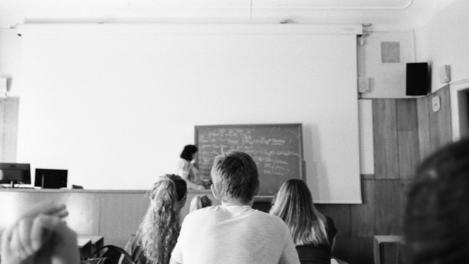 Skolēni klasē klausās skolotāju pie tāfeles melnbaltā fotogrāfijā