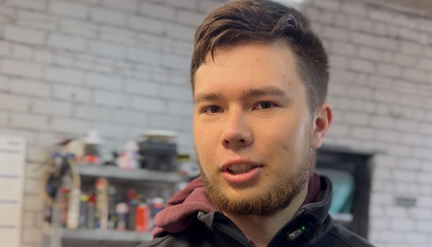 Jauns vīrietis stāv darbnīcā un runā kamerā; fonā redzami instrumenti un plaukti ar dažādiem materiāliem, radot amatniecības un radoša darba vidi