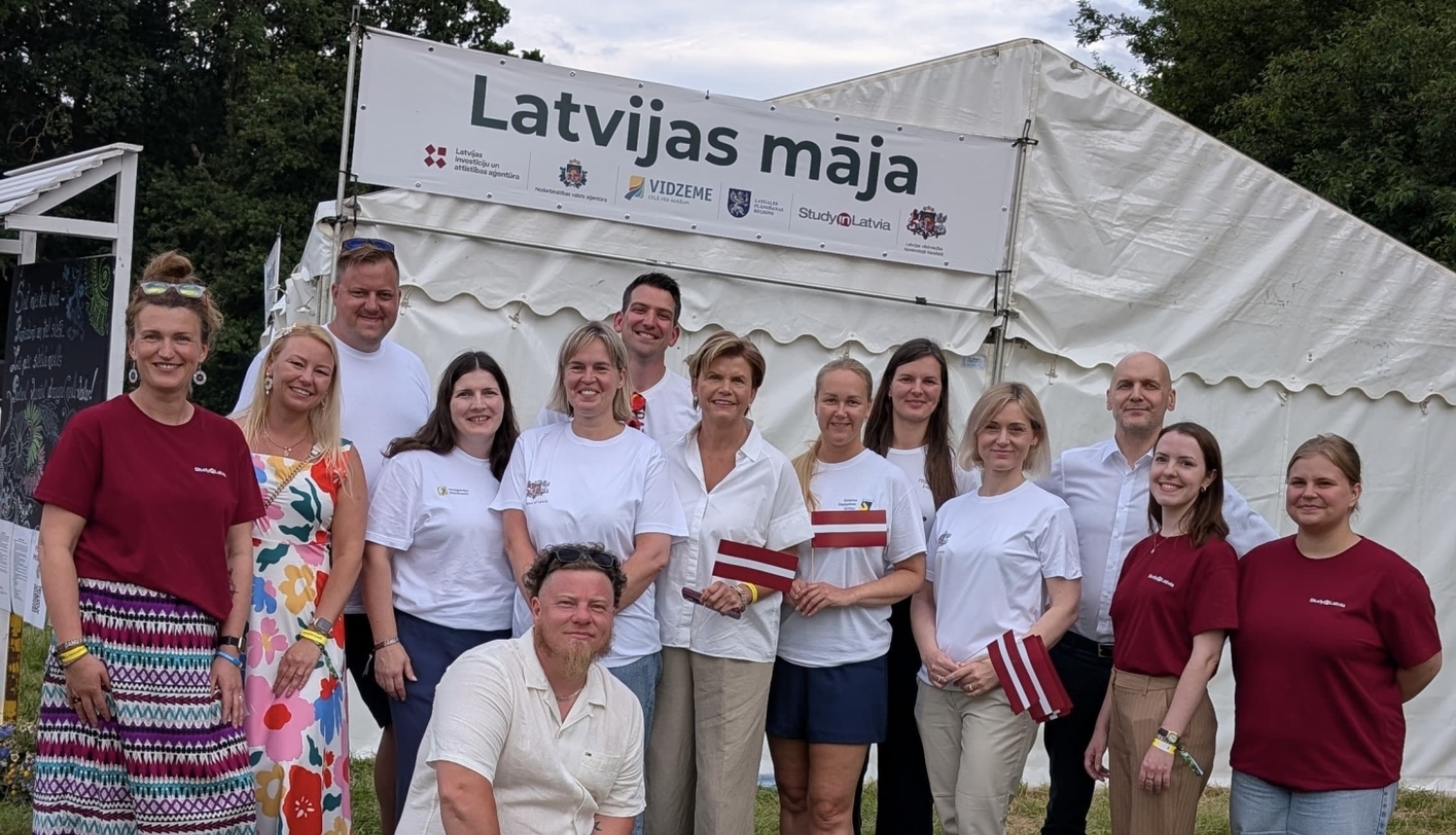 Cilvēku grupa pozē kopbildei pie “Latvijas mājas” telts āra festivālā