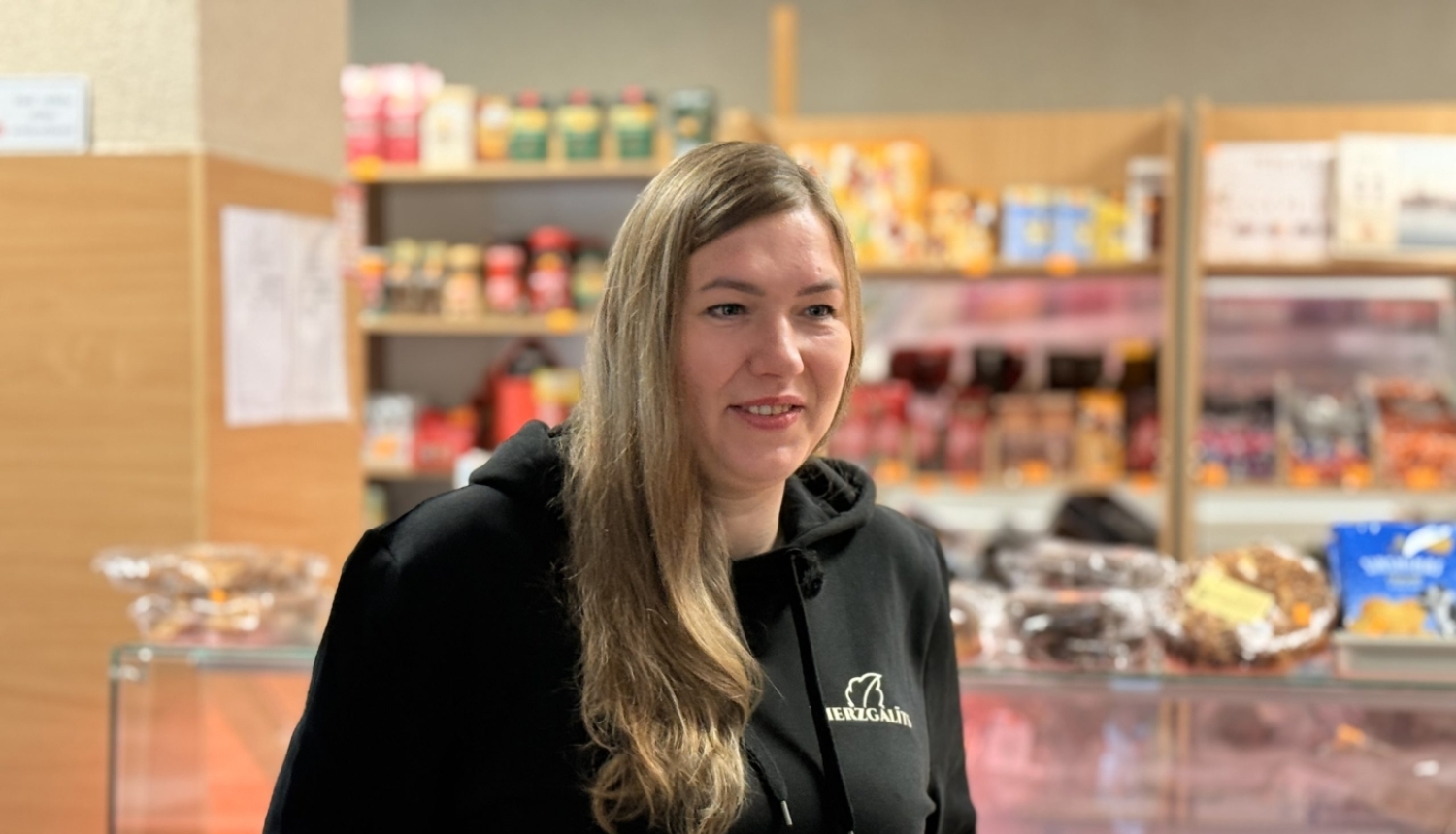 Sieviete melnā džemperī ar uzrakstu “Bierzgalīts” stāv lauku bodē pie vitrīnas ar pārtikas produktiem.