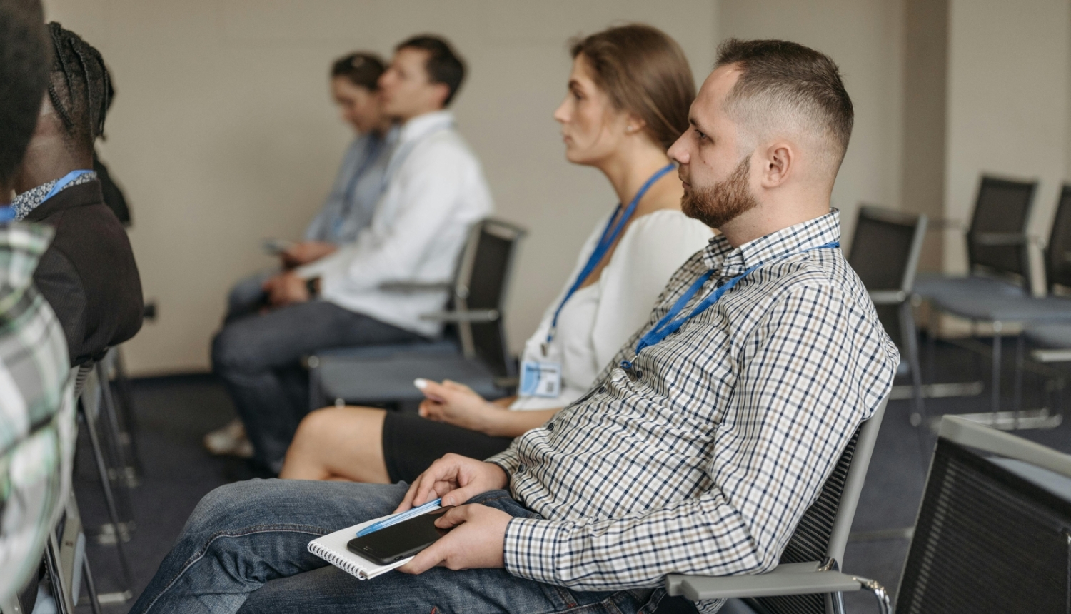 Cilvēki sēž seminārā vai konferencē, klausoties prezentāciju telpā