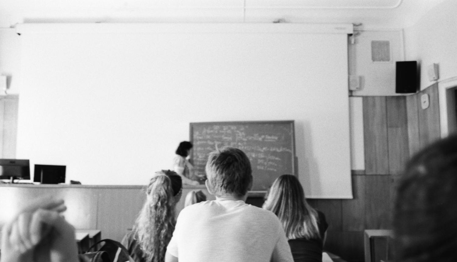 Skolēni klasē klausās skolotāju pie tāfeles melnbaltā fotogrāfijā