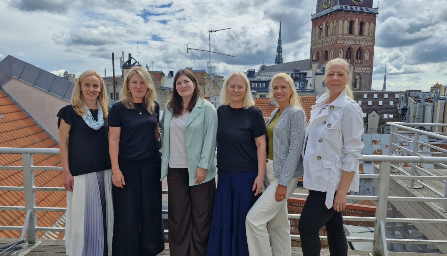 Six women stand on a terrace, posing for a photo. In the background, an old town skyline with a church spire and a clock tower is visible beneath a cloudy sky.